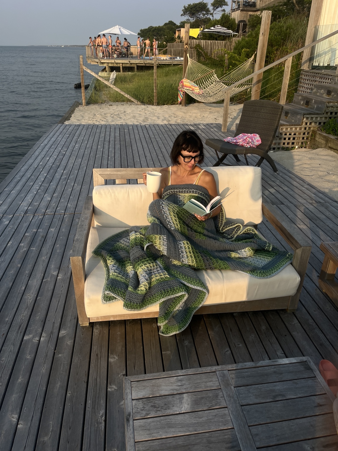 Home Handmade green and gray crochet blanket draped around a seated person reading with a mug on a Fire Island waterfront deck in golden light
